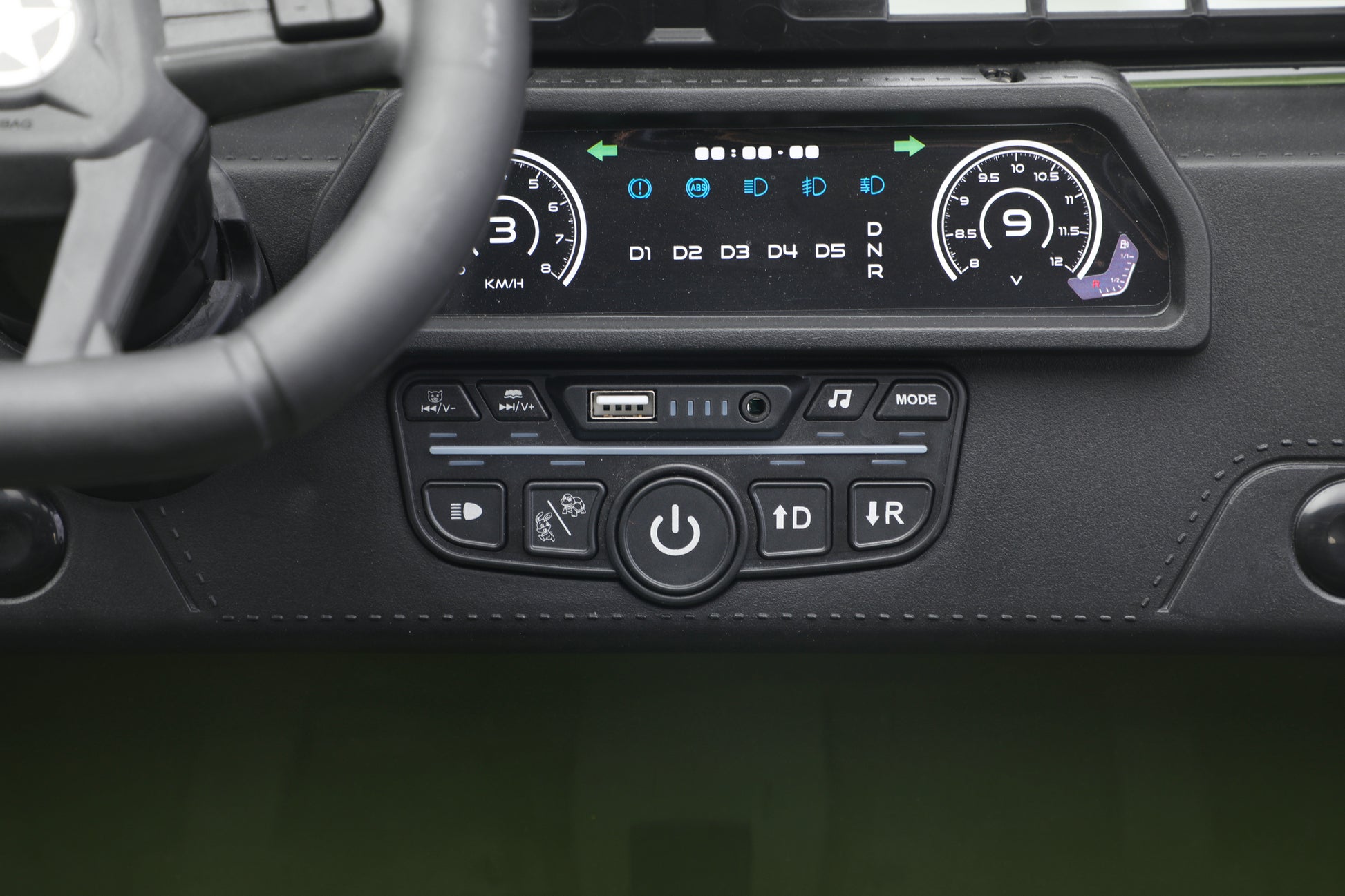Close-up of a toy jeep 24v car's dashboard centre console showing a digital instrument display and control buttons below, with part of the steering wheel visible on the left.